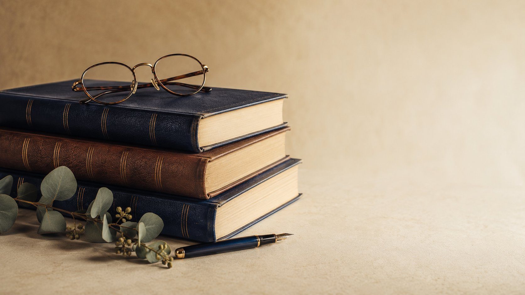 A stack of leather-bound books with reading glasses, a fountain pen and eucalyptus, in warm light