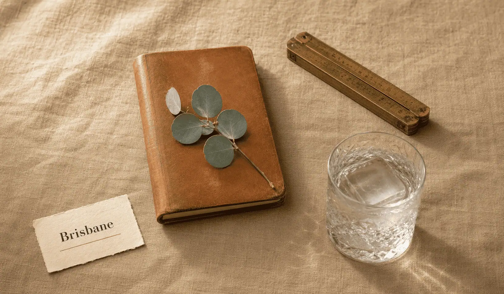 Brisbane anaesthetic billing service — editorial still life of a closed honey-tan leather logbook with a sprig of native silver-dollar eucalyptus resting on the cover, an etched crystal tumbler of water with one ice cube, a vintage brass folding ruler, and a small card reading Brisbane, on warm sandstone linen.